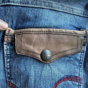May include: Close-up of a denim jeans pocket with a brown leather accent and a silver button that reads "BLUE TRIP". The jeans are a faded blue with red stitching.