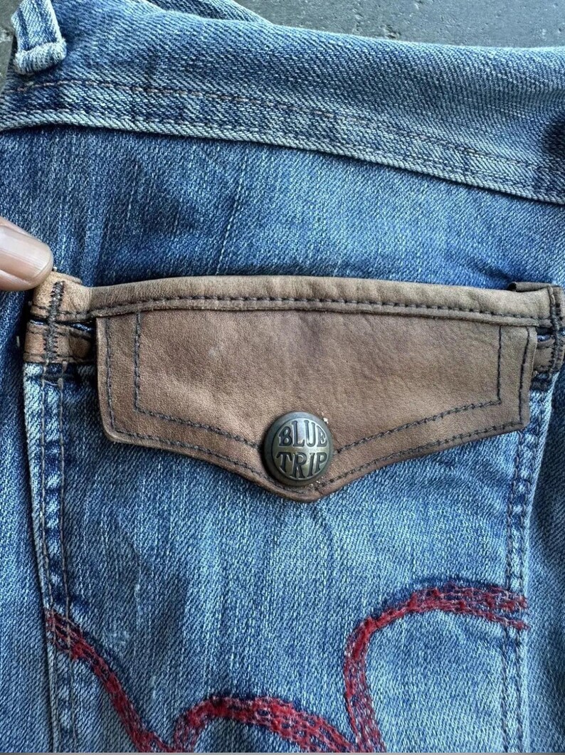 May include: Close-up of a denim jeans pocket with a brown leather accent and a silver button that reads "BLUE TRIP". The jeans are a faded blue with red stitching.