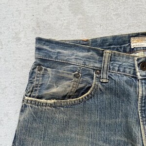 May include: Close-up of blue denim jeans with a faded, worn appearance. The jeans feature a classic five-pocket design, with visible stitching and rivets. The fabric shows signs of wear, adding to the vintage aesthetic. The jeans are a medium wash.