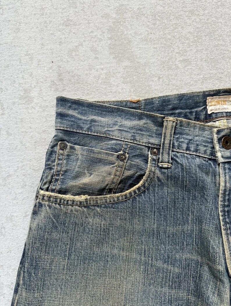 May include: Close-up of blue denim jeans with a faded, worn appearance. The jeans feature a classic five-pocket design, with visible stitching and rivets. The fabric shows signs of wear, adding to the vintage aesthetic. The jeans are a medium wash.