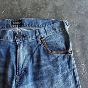 May include: Close-up of blue denim jeans with a faded wash. The jeans feature brown leather trim on the front pockets and a button closure. The brand name "BLUE TRIP" is visible on the waistband.