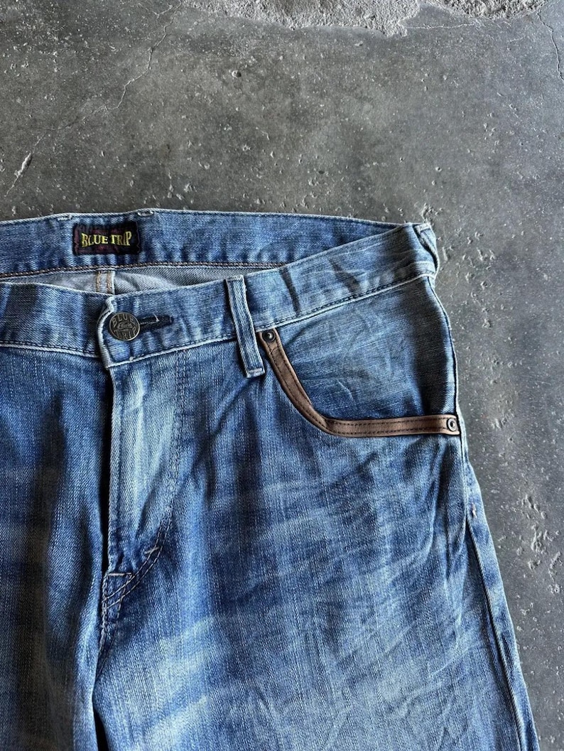 May include: Close-up of blue denim jeans with a faded wash. The jeans feature brown leather trim on the front pockets and a button closure. The brand name "BLUE TRIP" is visible on the waistband.