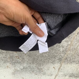 May include: Close-up of a black sweatshirt with a gray interior. The image shows the inside of the sweatshirt with a tag that has text in multiple languages. The sweatshirt is on a gray surface.
