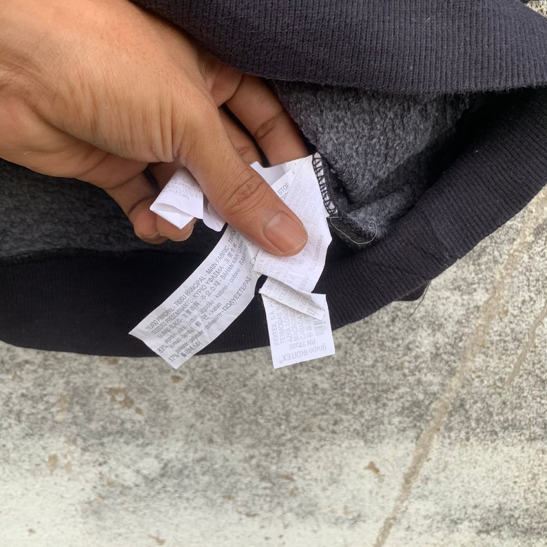 May include: Close-up of a black sweatshirt with a gray interior. The image shows the inside of the sweatshirt with a tag that has text in multiple languages. The sweatshirt is on a gray surface.