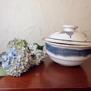 May include: A white and blue ceramic lidded bowl with a leaf design. The bowl has a blue band around the middle and sits on a dark wooden surface. A bouquet of blue and green hydrangeas is next to the bowl.