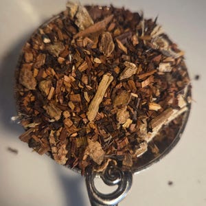May include: A metal measuring spoon filled with a loose-leaf tea blend consisting of small, dried brown and tan botanical pieces, including woody bark fragments, fibrous root segments, and dark tea leaves, all piled together in the round bowl.