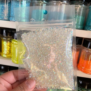 May include: A clear plastic bag filled with iridescent white round beads. The bag is sealed and has a hole at the top for hanging.