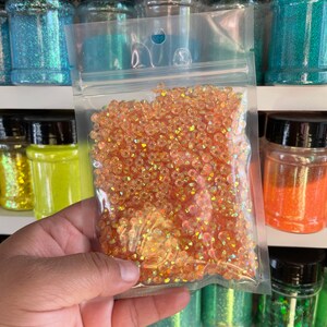 May include: A clear plastic bag filled with iridescent orange glitter. The glitter is in a variety of shapes and sizes. The bag is sitting on a shelf with other jars of glitter.