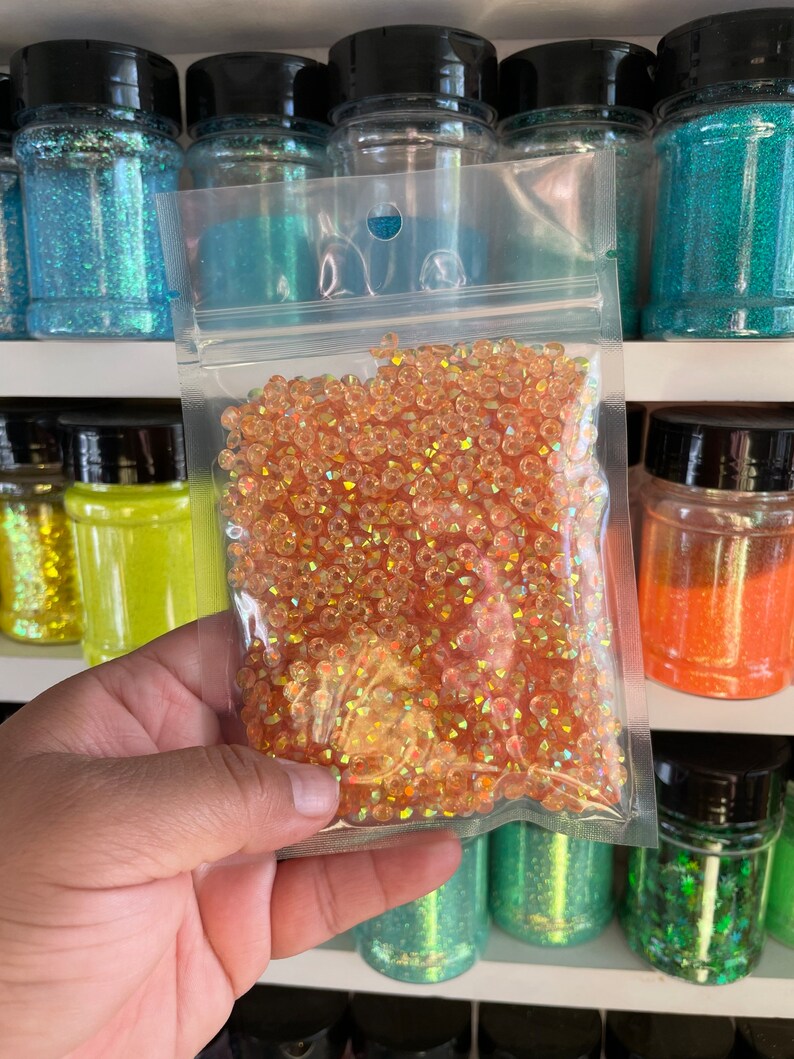 May include: A clear plastic bag filled with iridescent orange glitter. The glitter is in a variety of shapes and sizes. The bag is sitting on a shelf with other jars of glitter.