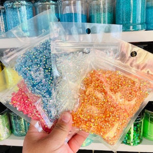May include: Four bags of iridescent glass beads in different colors: light blue, clear, orange, and pink. The bags are held in a hand.
