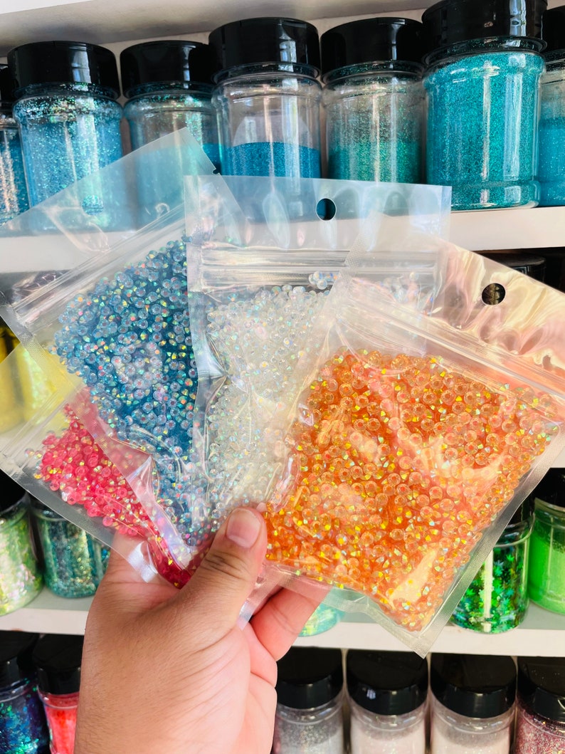 May include: Four bags of iridescent glass beads in different colors: light blue, clear, orange, and pink. The bags are held in a hand.