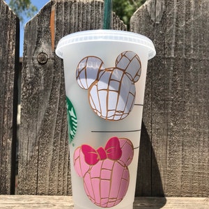 May include: A clear plastic reusable cup with a straw, featuring a pink and white Mickey Mouse head design with a pink bow. The design is inspired by a Mexican pastry called a concha.