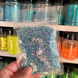 May include: A clear plastic bag filled with iridescent blue glitter. The bag is held in a hand and is in front of a shelf of jars filled with various colors of glitter.