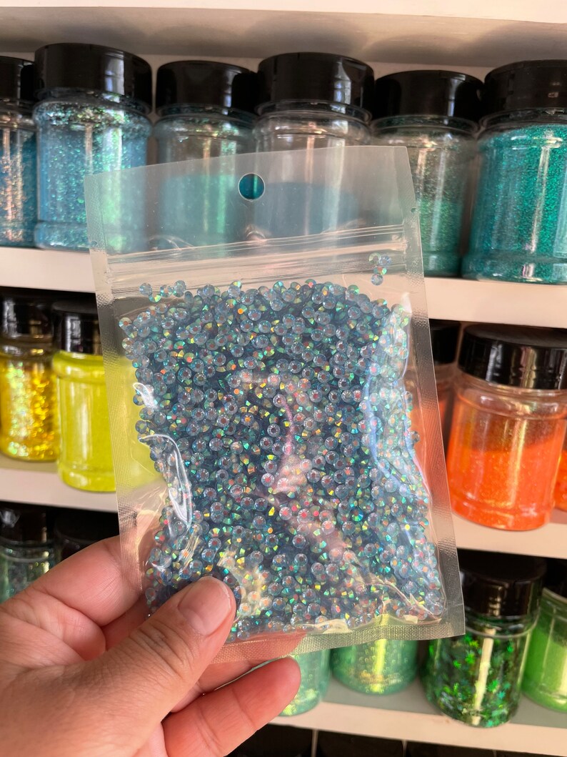 May include: A clear plastic bag filled with iridescent blue glitter. The bag is held in a hand and is in front of a shelf of jars filled with various colors of glitter.