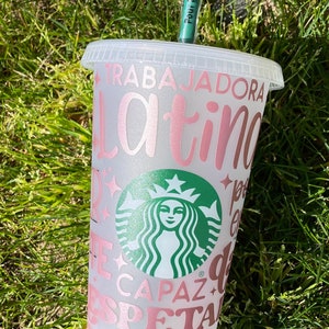 May include: A white reusable cup with a pink and brown design. The cup has the text "Trabajadora Latina" and a Starbucks logo. The cup is sitting on a bed of green grass.