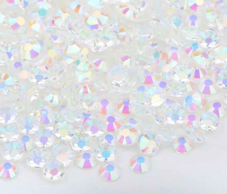 May include: A pile of iridescent clear glass rhinestones. The rhinestones are round and have a faceted design.