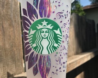 May include: A clear, reusable cold cup with a green Starbucks logo. The cup is decorated with a purple sunflower design and musical notes. The sunflower petals have a holographic, iridescent finish.