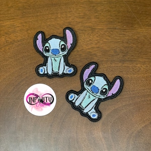 May include: Two blue and purple embroidered iron-on patches of Stitch from Lilo & Stitch. The patches are on a brown wooden surface.