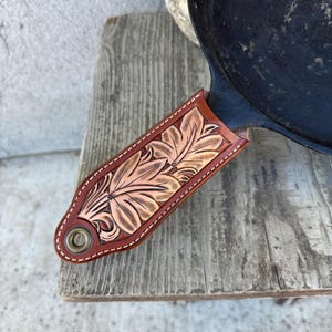 May include: A cast iron skillet with a decorative, hand-tooled leather handle cover. The handle cover is brown with a floral design in a lighter brown color. The skillet is black and sits on a weathered wooden surface.