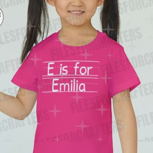 Puede incluir: Camiseta rosa con texto blanco que dice "E es para Emilia".