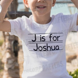 Puede incluir: Una camiseta blanca con texto negro que dice "J es para Joshua" sobre un fondo rayado. La camiseta la lleva puesta un ni&ntilde;o.