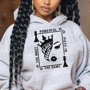 Puede incluir: Sudadera blanca con un dise&ntilde;o gr&aacute;fico negro que presenta una pieza de ajedrez de reina con una corona y el texto "The Most Powerful Piece In The Game" y "Black Queen".