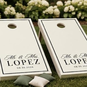 Könnte beinhalten: Zwei weiße Cornhole-Boards mit schwarzen rechteckigen Rändern und kreisförmigen Löchern an der Oberseite. Jedes Board zeigt den schwarzen Text Mr. & Mrs. LOPEZ und das Datum 08.30.2026. Drei kleine Sitzsäcke in Olivgrün und Weiß liegen davor.