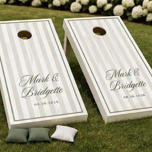 Könnte beinhalten: Zwei weiße Cornhole-Boards mit vertikalen hellgrauen Streifen und einem kreisförmigen Loch an der Oberseite. Die Boards sind mit dem Text Mark & Bridgette und dem Datum 08.30.2026 in einer dunkelgrauen Schreibschrift personalisiert. Drei Säckchen liegen bei.