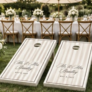 Könnte beinhalten: Zwei rechteckige Cornhole-Boards mit einem beigefarbenen und weißen vertikalen Streifenmuster und einem dunklen Rand. Jedes Board zeigt den Text Mr. & Mrs. Bradley und das Datum MAY 8, 2027 in schwarzer Schreib- und Serifenschrift mit einem Loch.