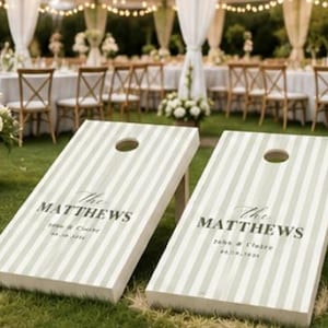 Könnte beinhalten: Zwei hölzerne Cornhole-Boards mit einem vertikalen Streifenmuster in hellem Salbeigrün und Weiß. Jedes Board zeigt den Text The MATTHEWS, John & Claire und 06.20.2024 in einer dunkelgrauen Serifenschrift sowie ein rundes Loch.