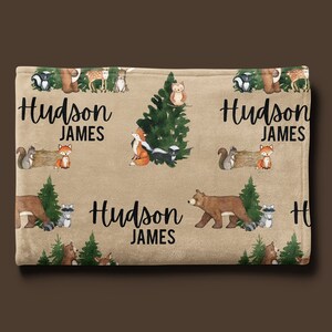 Puede incluir: Una manta de franela marr&oacute;n y beige con estampado de animales del bosque. La manta presenta un patr&oacute;n repetido de osos, zorros, mapaches, ardillas, b&uacute;hos y ciervos. La manta est&aacute; personalizada con el nombre "Hudson James" repetido en toda la superficie.