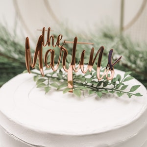 May include: A white cake with a rose gold cake topper that says "the Martins".