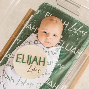 May include: A newborn baby wrapped in a white swaddle blanket, lying on a green blanket inside a clear bassinet. The green blanket has the name "Elijah Lukas" printed on it. A wooden round sign with the name "Elijah" in green letters is placed on the blanket.