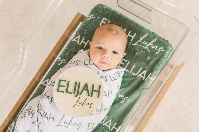 May include: A newborn baby wrapped in a white swaddle blanket, lying on a green blanket inside a clear bassinet. The green blanket has the name "Elijah Lukas" printed on it. A wooden round sign with the name "Elijah" in green letters is placed on the blanket.