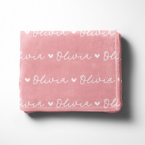 May include: A pink fleece blanket with the name "Olivia" repeated in white script with small white hearts between each name.
