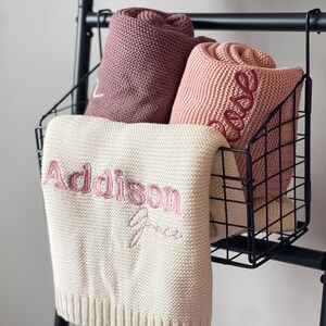K&ouml;nnte beinhalten: Drei gestrickte Decken in einem Drahtkorb. Die Decken sind rosa, hellrosa und wei&szlig;. Die wei&szlig;e Decke hat den Namen "Addison Grace" in rosa Garn gestickt.