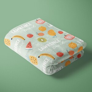 May include: A soft, light blue blanket with a colorful fruit pattern. The blanket features a repeating pattern of bananas, strawberries, pineapples, watermelons, apples, and pears. The text "LAWRENCE lived" is repeated throughout the blanket.