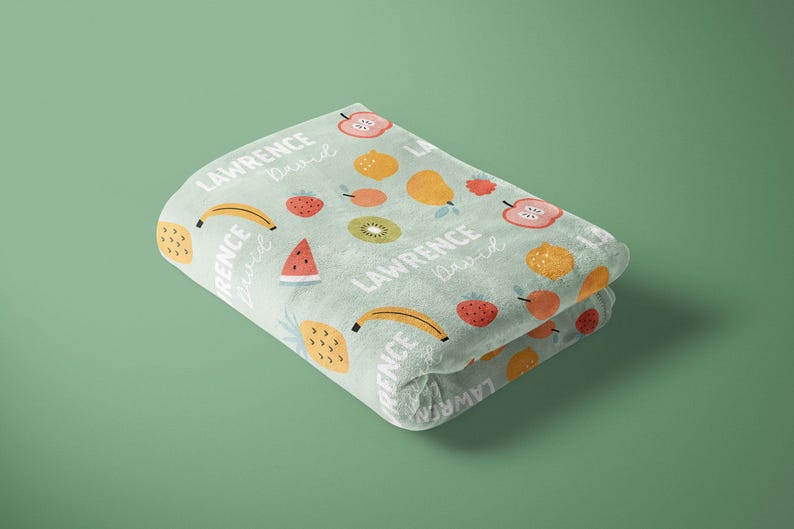 May include: A soft, light blue blanket with a colorful fruit pattern. The blanket features a repeating pattern of bananas, strawberries, pineapples, watermelons, apples, and pears. The text "LAWRENCE lived" is repeated throughout the blanket.