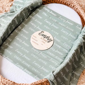 May include: A sage green baby blanket with the name "Emery Jeanne" repeated in white text. A wooden baby announcement plaque with the name "Emery Jeanne" and spaces for date, weight, time, length, lbs, and oz.