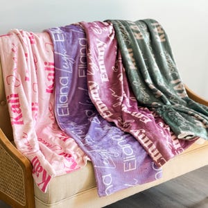May include: Four personalized blankets in shades of pink, purple, and green are draped over a beige sofa. Each blanket features a different name in a cursive font. The blankets are soft and plush, perfect for snuggling.