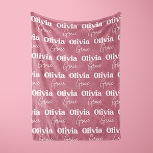 May include: A dusty rose-colored blanket with the names "Olivia" and "Grace" repeated in a white, stylized font. The blanket is hanging against a matching pink background, creating a soft and inviting aesthetic.