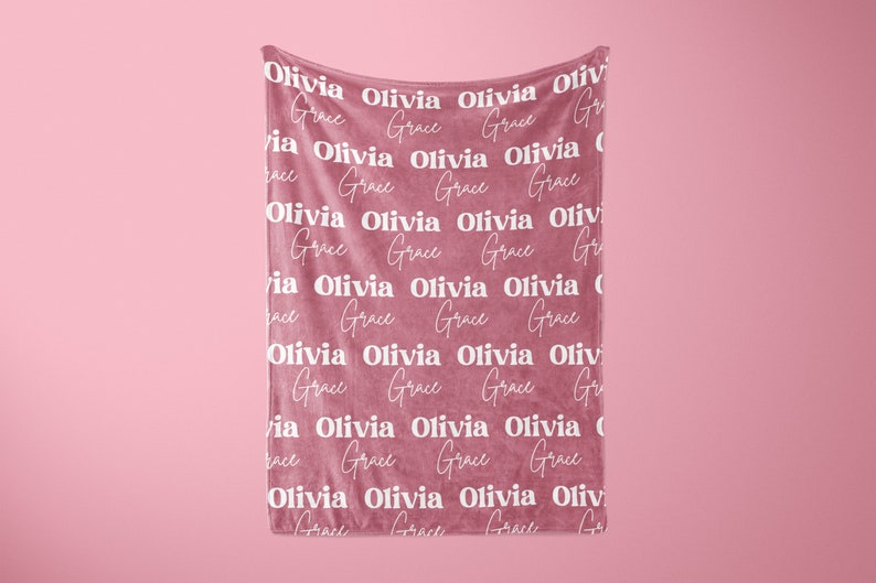 May include: A dusty rose-colored blanket with the names "Olivia" and "Grace" repeated in a white, stylized font. The blanket is hanging against a matching pink background, creating a soft and inviting aesthetic.