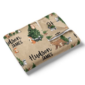 Puede incluir: Una manta para beb&eacute; de color beige y blanco con un estampado de animales del bosque. La manta presenta un oso, un zorro, un mapache, una mofeta y un b&uacute;ho. La manta est&aacute; personalizada con el nombre "Hudson James".