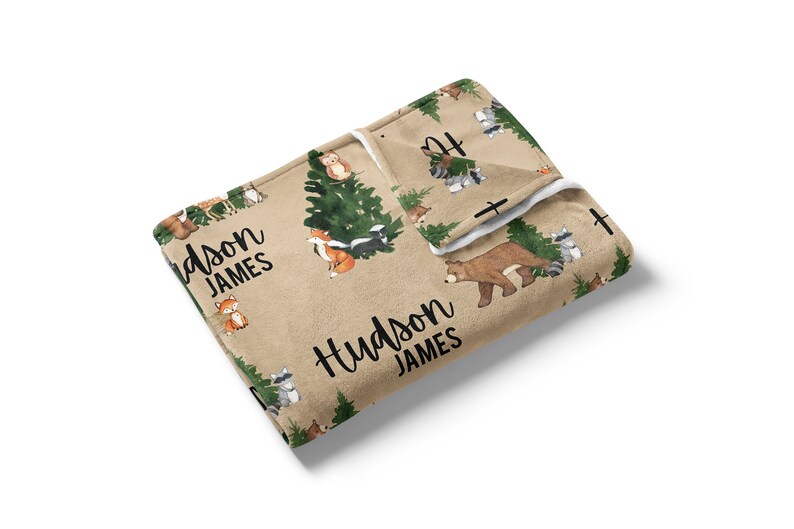 Puede incluir: Una manta para beb&eacute; de color beige y blanco con un estampado de animales del bosque. La manta presenta un oso, un zorro, un mapache, una mofeta y un b&uacute;ho. La manta est&aacute; personalizada con el nombre "Hudson James".