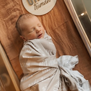 May include: A newborn baby is swaddled in a gray blanket with the words "Oaks Truman" printed on it. The baby is sleeping peacefully in a bassinet with a wooden sign that reads "Oaks Truman" above it.