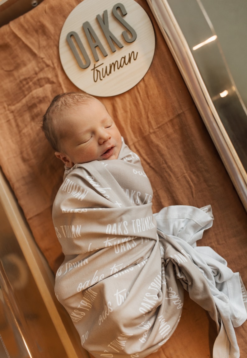 May include: A newborn baby is swaddled in a gray blanket with the words "Oaks Truman" printed on it. The baby is sleeping peacefully in a bassinet with a wooden sign that reads "Oaks Truman" above it.