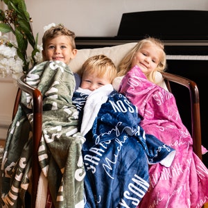 May include: Three children are sitting in a chair, each wrapped in a personalized fleece blanket. The blankets are green, blue, and pink, with white lettering. The children are smiling and looking at the camera.