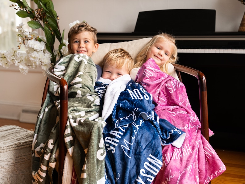 May include: Three children are sitting in a chair, each wrapped in a personalized fleece blanket. The blankets are green, blue, and pink, with white lettering. The children are smiling and looking at the camera.