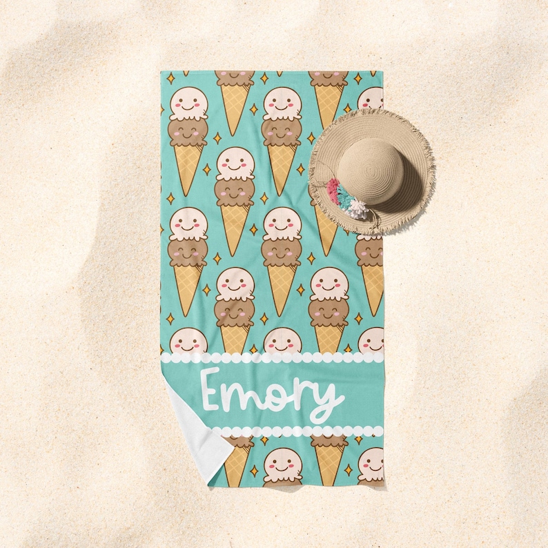 May include: A turquoise beach towel with a pattern of ice cream cones with smiling faces. The towel has a white border with the name "Emory" written in white.