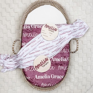 May include: A baby gift set featuring a woven basket, a plush blanket, and two wooden name plaques. The blanket and plaques display the name "Amelia Grace" in a cursive font. The blanket is white with the name in a mauve color, and the basket is lined with a mauve blanket.